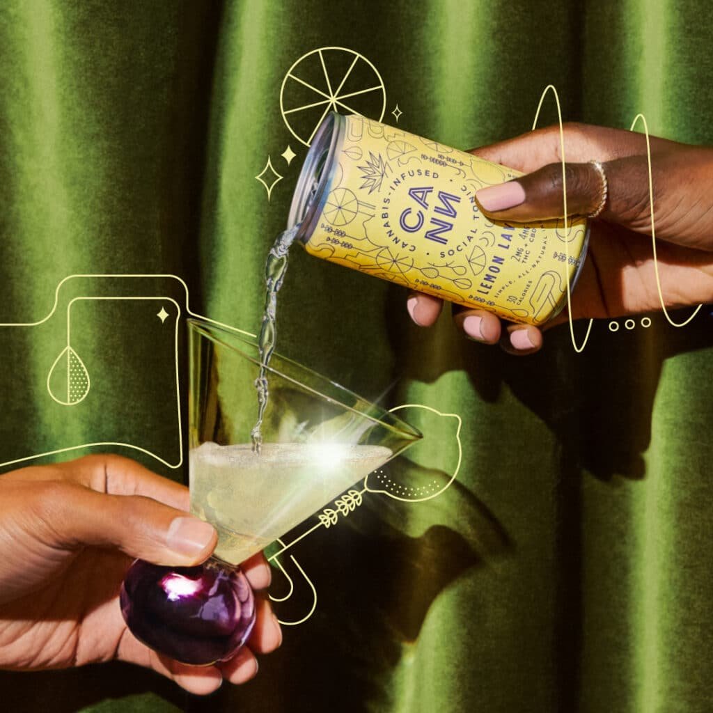 One hand pours a can of Lemon Lavender Cann into a martini-style glass that is being held at an angle by another hand in front of a green curtain background.
