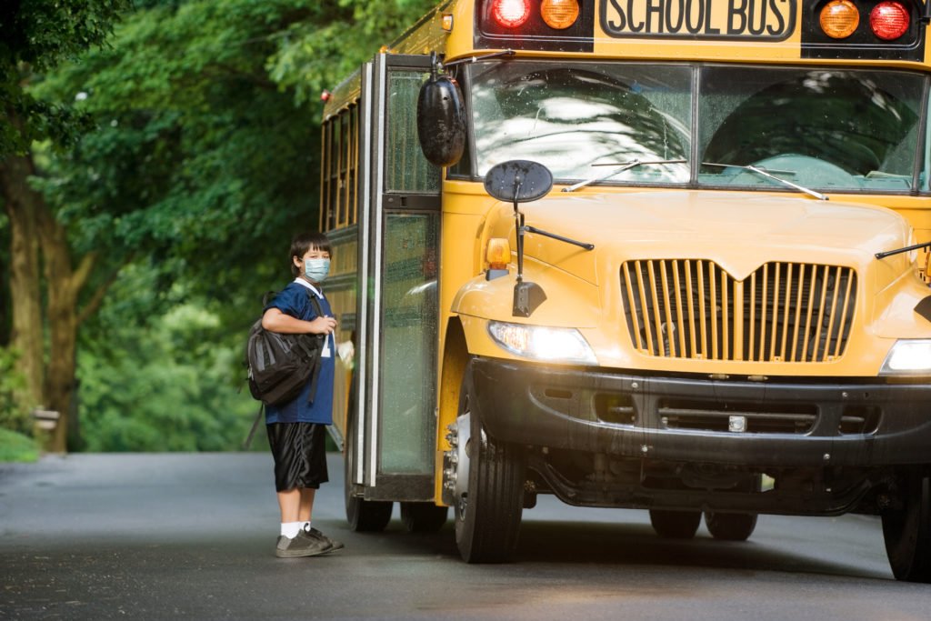 School bus picking up elementary student wearing surgical mask boarding at bus stop.