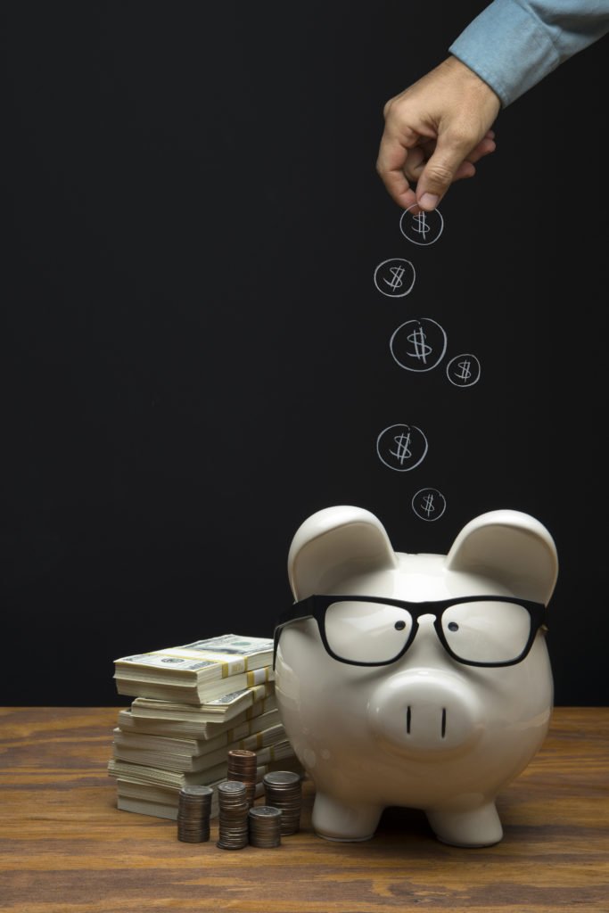 This is a photograph of a large white Piggy Bank sitting on a desk a with a male hand depositing a chalk drawing of money coins on Black Chalkboard Background. This image could relate to savings and retirement.