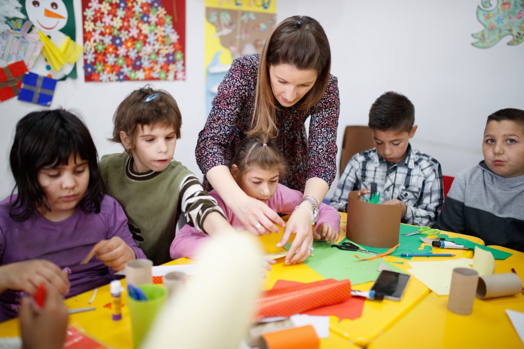 Special education teacher standing by the desk with paper craft in art class and assisting elementary students