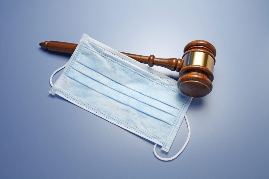 A gavel rests on top of a protective mask on a blue background. The image conveys the concept legal issues relating to COVID-19.