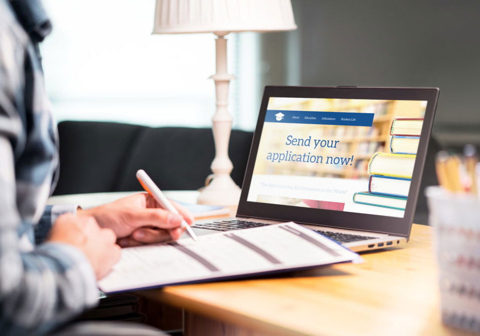 Young man writing college or university application form with pen and paper. Student applying or filling document. Scholarship, grant or admission. School website in laptop.