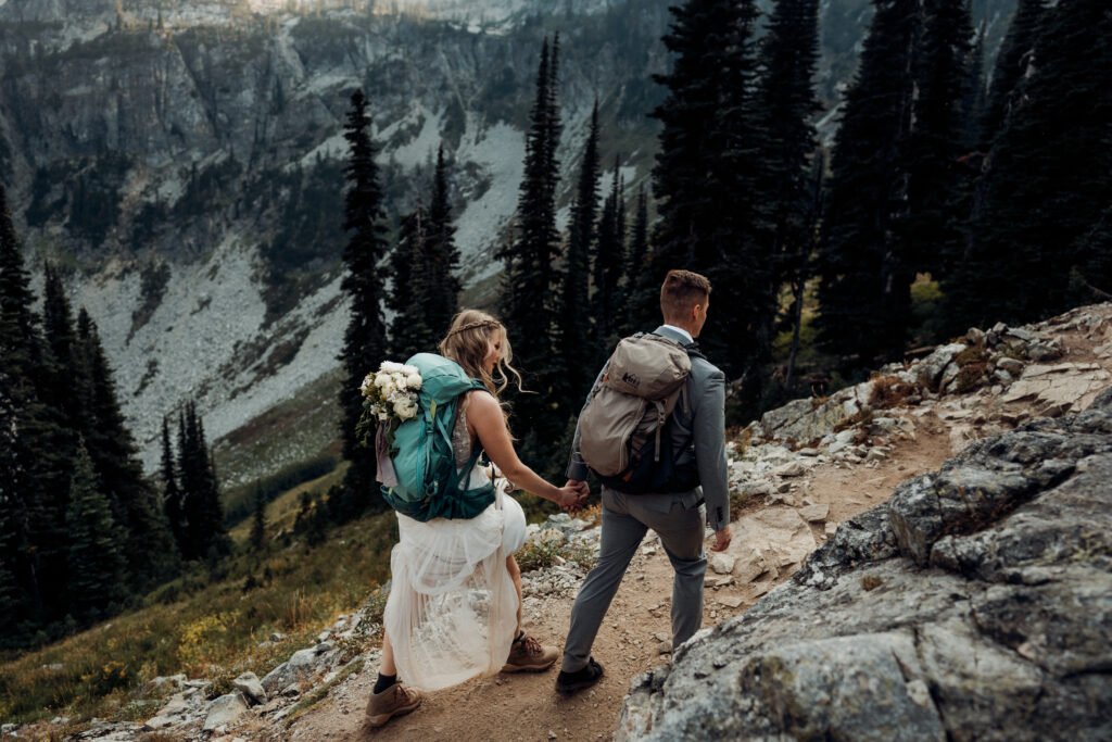 A couple holds has they hike a trail together in the mountains in their wedding attire. 