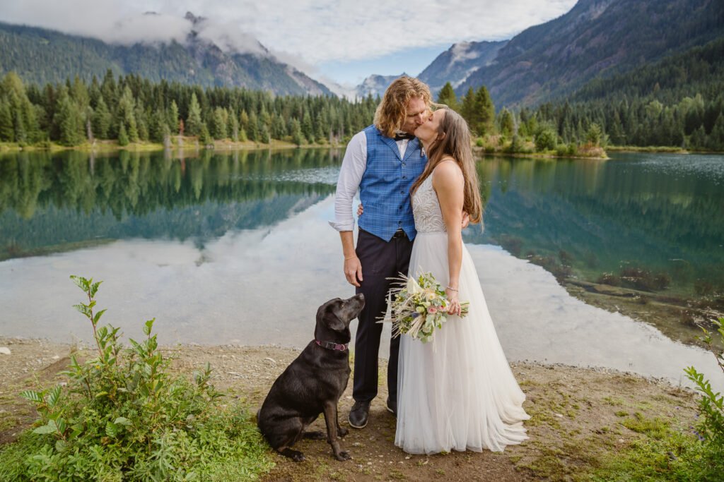 A couple shares a kiss in the mountains as their dog looks on.